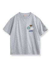 左胸に4つのカラフルな刺繍バッジが付いたグレーベースの半袖Tシャツ。