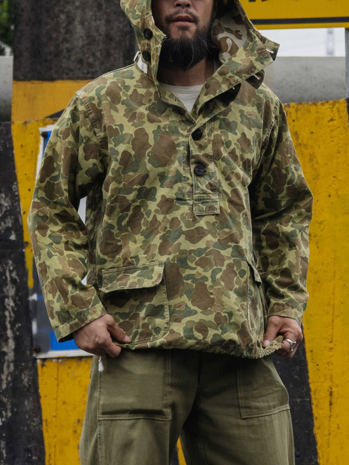 詳細画像: 大きなフードと三層前立てデザインのディテール特写。機能性とヴィンテージ感が際立つディテール。
モデル画像: モデルが迷彩柄砲兵防護プルオーバーパーカーを着用したスタイリング例。戦時風デザインを取り入れたコーディネート。