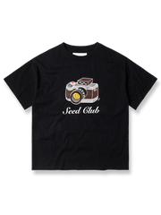 製品画像: コンリホワイトカメラプリントTシャツの全体像、クラシックなカメラモチーフとルーズフィットシルエットを示す。