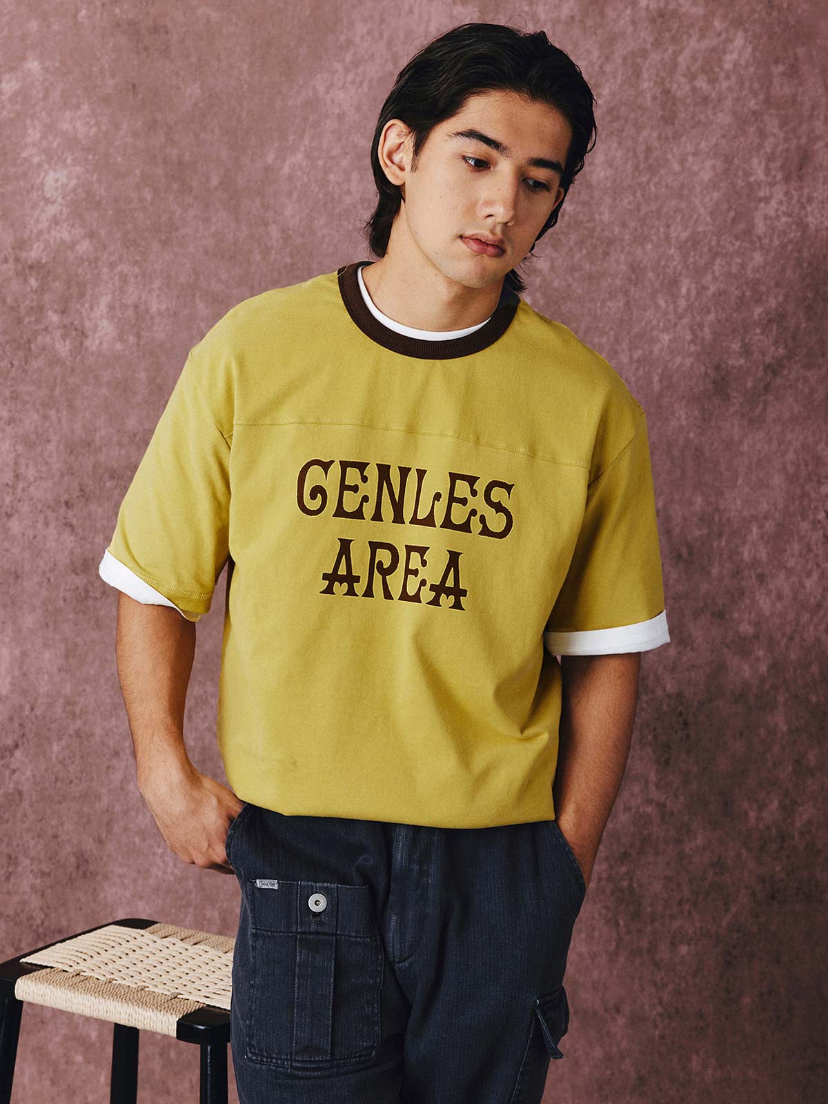 モデルがフットボールカルチャープリントTシャツを着用し、ジンジャーイエローのバリエーションで、ダークカラーのカジュアルパンツとスポーツシューズを合わせたカジュアルなスタイリングでポーズをとる画像。レトロかつ現代的な夏の装いを演出。