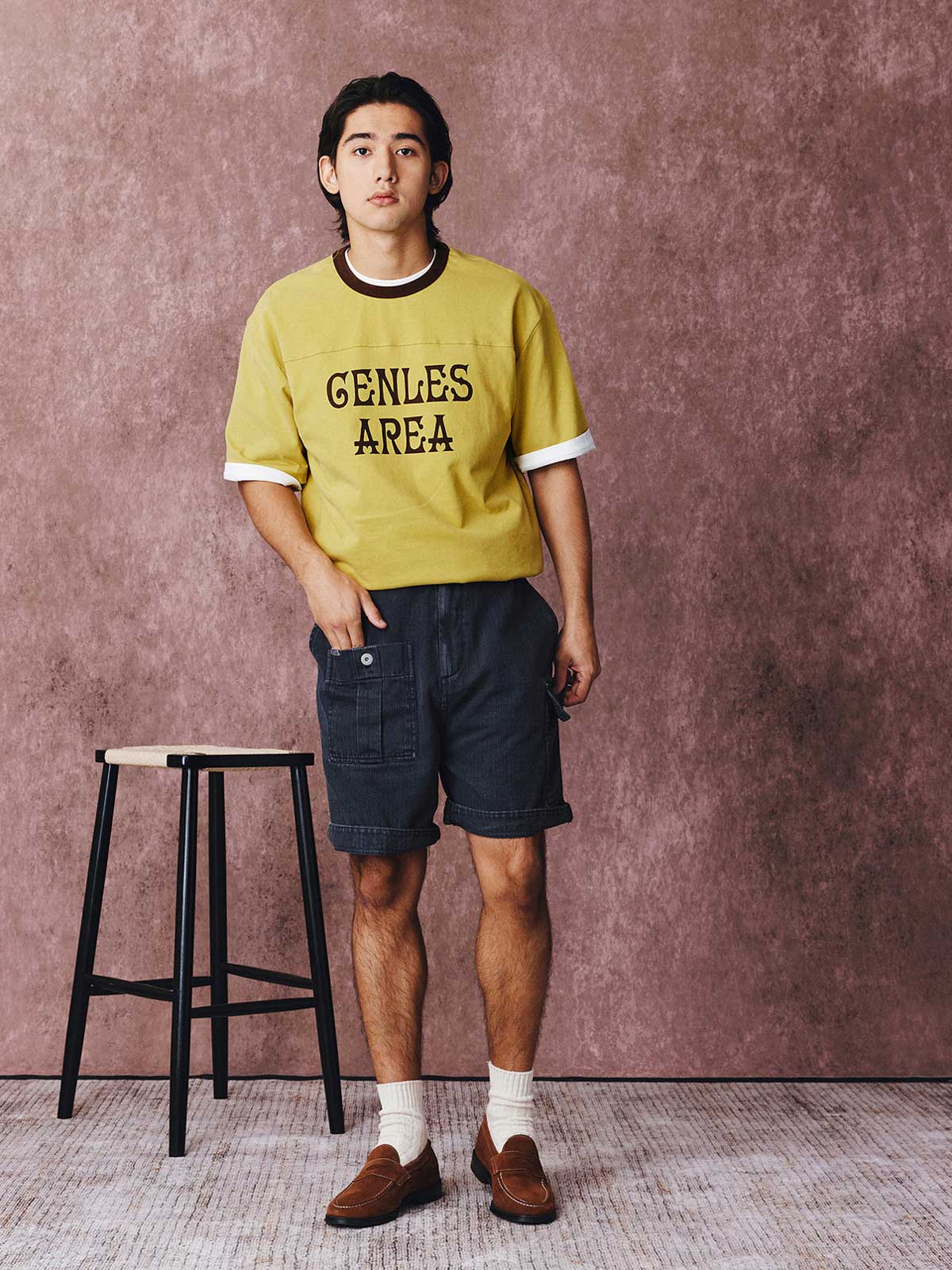 モデルがフットボールカルチャープリントTシャツを着用し、ジンジャーイエローのバリエーションで、ダークカラーのカジュアルパンツとスポーツシューズを合わせたカジュアルなスタイリングでポーズをとる画像。レトロかつ現代的な夏の装いを演出。