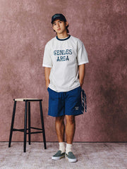 モデルがフットボールカルチャープリントTシャツを着用し、ジンジャーイエローのバリエーションで、ダークカラーのカジュアルパンツとスポーツシューズを合わせたカジュアルなスタイリングでポーズをとる画像。レトロかつ現代的な夏の装いを演出。