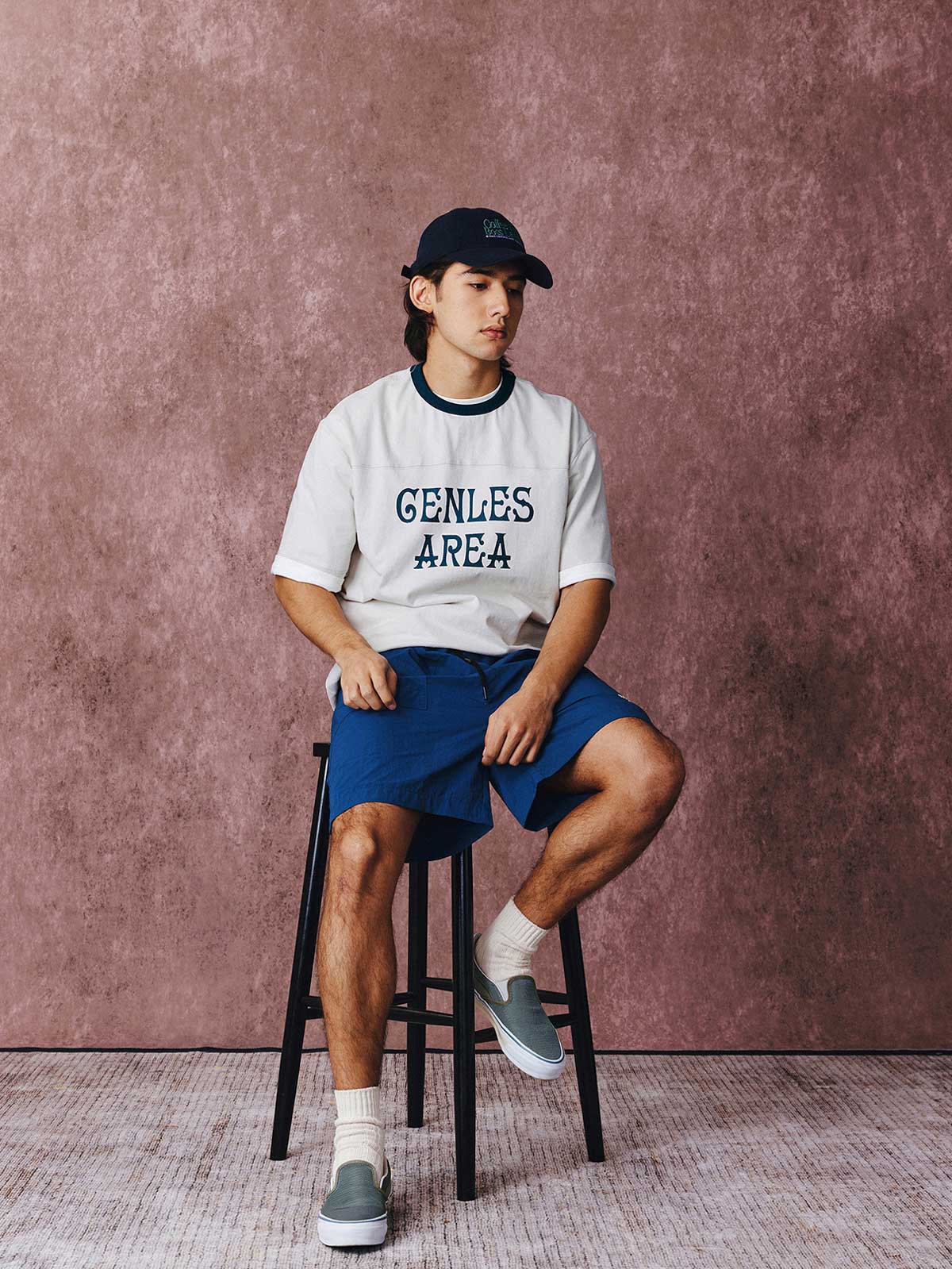 モデルがフットボールカルチャープリントTシャツを着用し、ジンジャーイエローのバリエーションで、ダークカラーのカジュアルパンツとスポーツシューズを合わせたカジュアルなスタイリングでポーズをとる画像。レトロかつ現代的な夏の装いを演出。