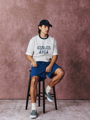 モデルがフットボールカルチャープリントTシャツを着用し、ジンジャーイエローのバリエーションで、ダークカラーのカジュアルパンツとスポーツシューズを合わせたカジュアルなスタイリングでポーズをとる画像。レトロかつ現代的な夏の装いを演出。