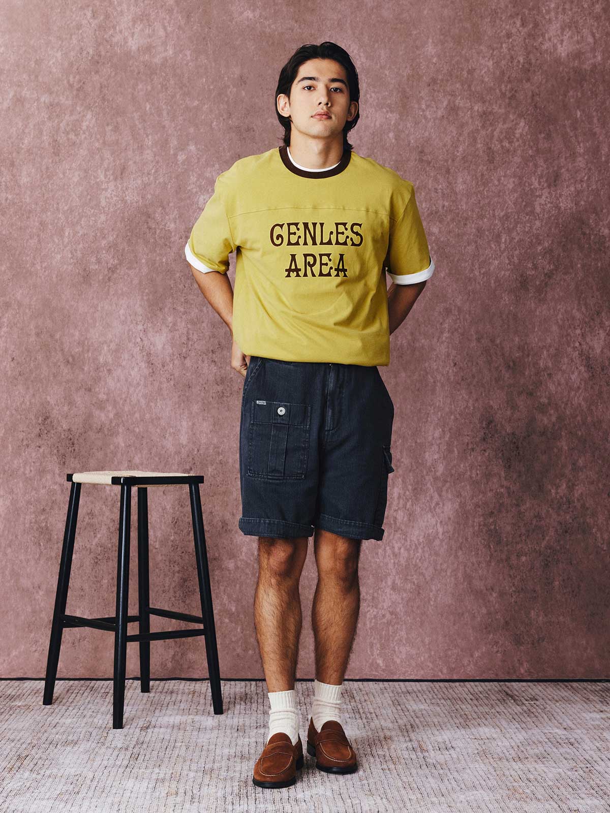 モデルがフットボールカルチャープリントTシャツを着用し、ジンジャーイエローのバリエーションで、ダークカラーのカジュアルパンツとスポーツシューズを合わせたカジュアルなスタイリングでポーズをとる画像。レトロかつ現代的な夏の装いを演出。