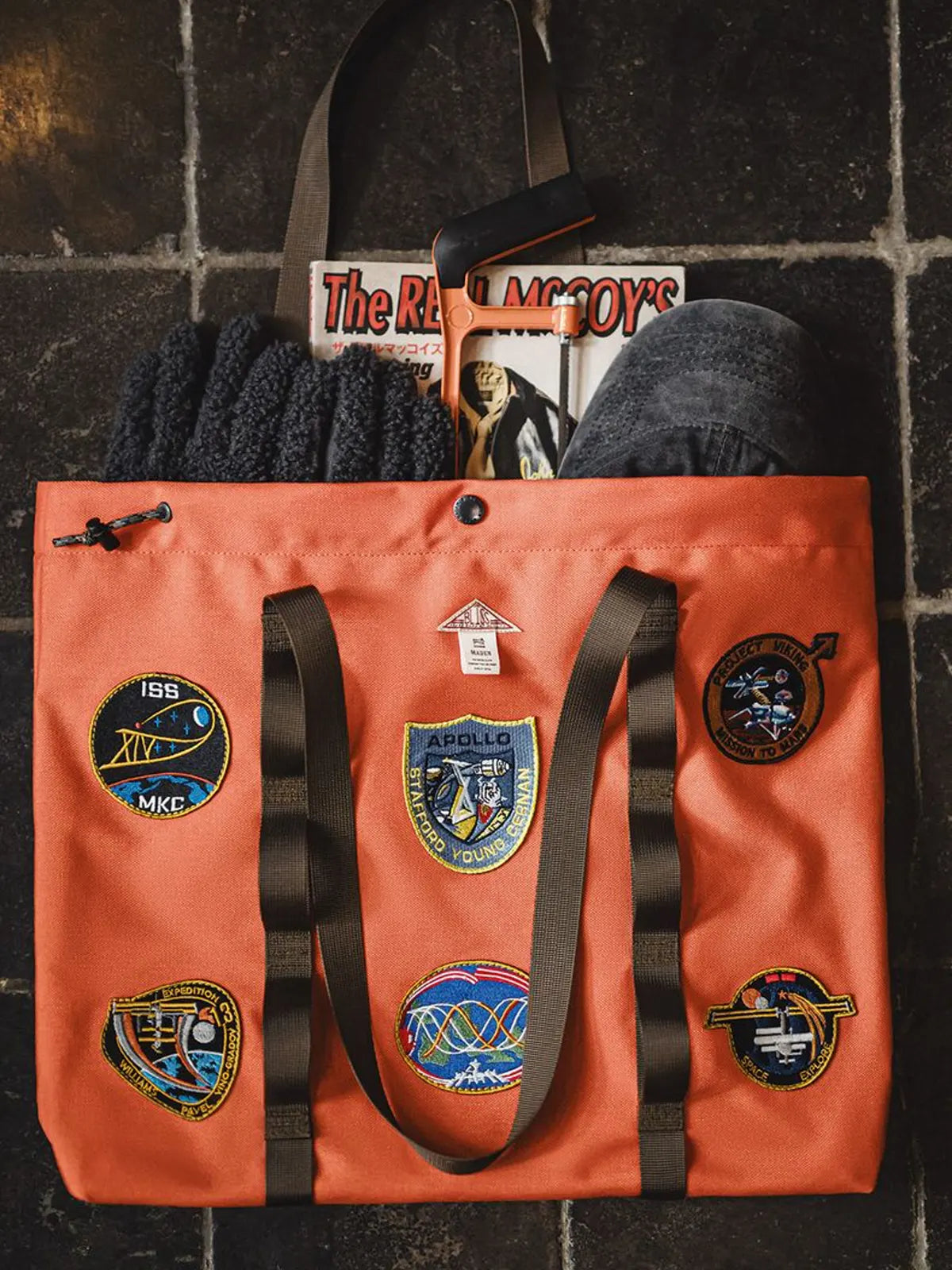 アポロ号テーマの刺繍が施されたオレンジ色のナイロン製トートバッグ。大容量で機能性があり、デザイン性も高い。

