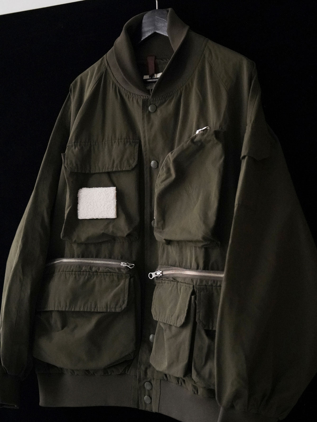 製品画像
「アシンメトリーデザインの多ポケットキャンプワークジャケット全体の展示図。クラシックなフィッシングジャケットとワークスタイルが融合したデザインで、ユニークなアシンメトリーデザインが実用性と個性を強調。防撥水機能を備えたポリエステルとコットン混紡生地を使用し、快適な着心地を提供します。」

詳細画像
「胸元のロゴ刺繍とアシンメトリーな多機能ポケットのディテール特写。YKKカスタムジッパーとロゴ入りスナップボタンが、高品質のディテールを引き立て、機能性とスタイルを両立させています。ダブルレイヤーのリブ編みが施され、袖口と裾のフィット感と耐久性を向上させています。」
