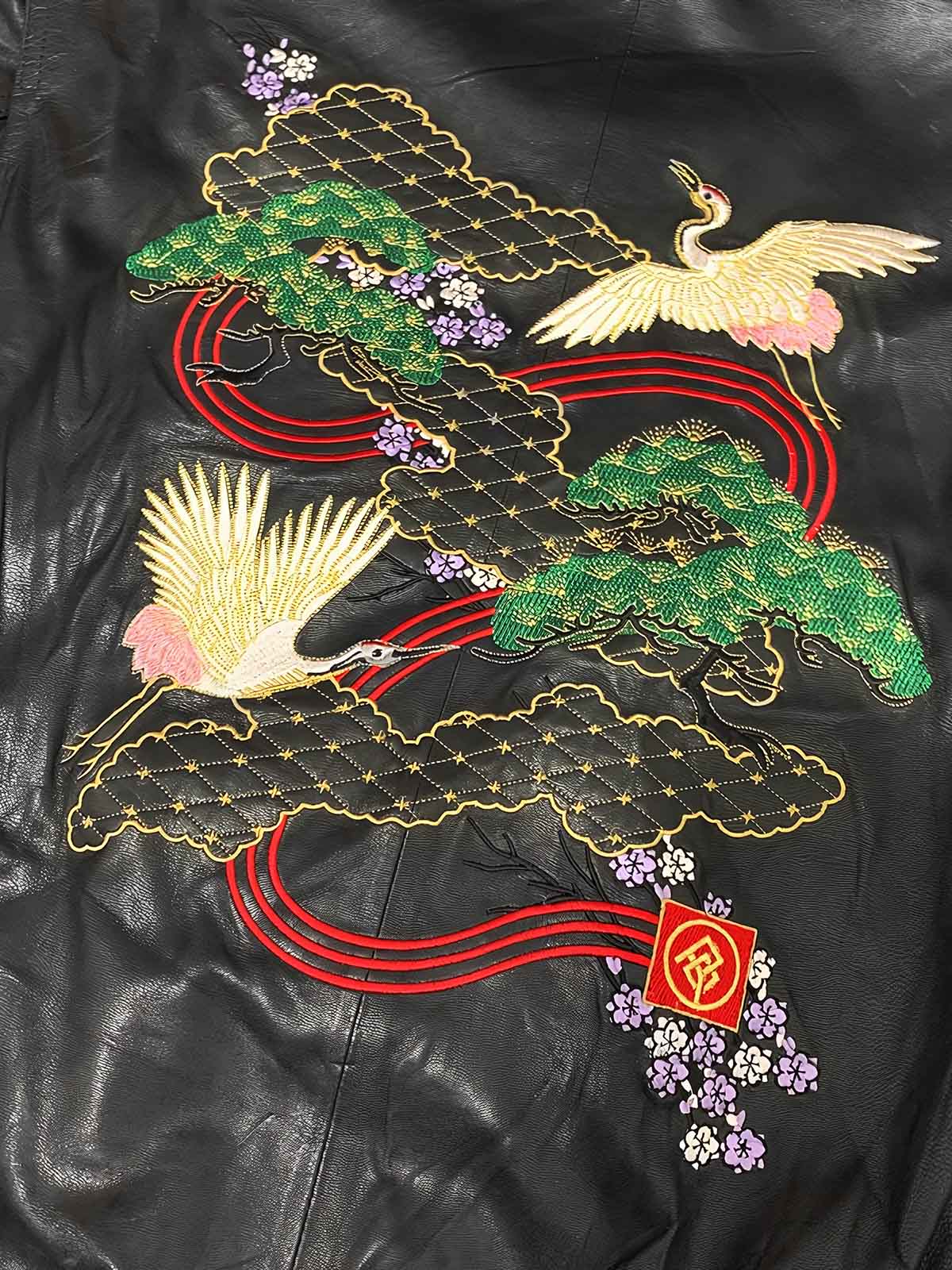 製品画像: 横須賀鶴図刺繍 羊革ジャケット全体画像、吉祥の象徴である「鶴図」刺繍と上質な輸入羊革のデザイン
詳細画像: 横須賀の伝統技法を用いた鶴、松、雲の刺繍ディテール、立体感ある精巧な工芸