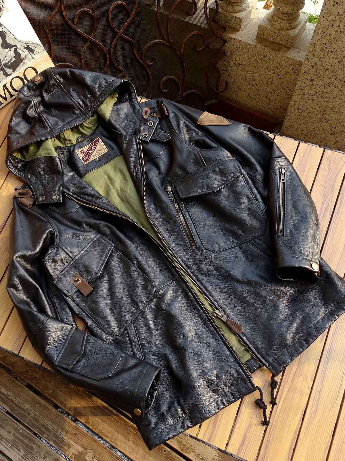 製品画像： 9ポケットフィールドフード付きフルグレインレザージャケット全体イメージ画像。黒いシボ加工レザーとヴィンテージ感漂うオイル仕上げのスエードディテールが特徴で、アウトドアやバイクライドにも適した多機能デザイン。
詳細画像： YKKアンティークブロンズジッパーとオイルドスエードレザーの補強ディテールをクローズアップした画像。耐久性とクラシックな風合いが際立つ仕上がり。