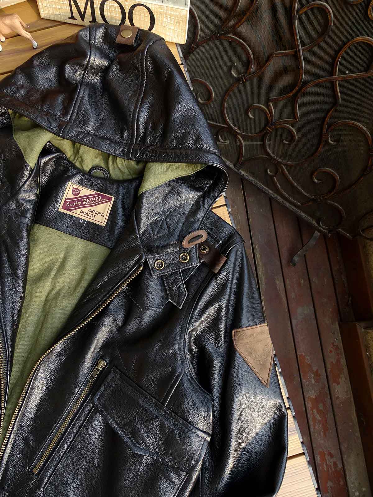 製品画像： 9ポケットフィールドフード付きフルグレインレザージャケット全体イメージ画像。黒いシボ加工レザーとヴィンテージ感漂うオイル仕上げのスエードディテールが特徴で、アウトドアやバイクライドにも適した多機能デザイン。
詳細画像： YKKアンティークブロンズジッパーとオイルドスエードレザーの補強ディテールをクローズアップした画像。耐久性とクラシックな風合いが際立つ仕上がり。