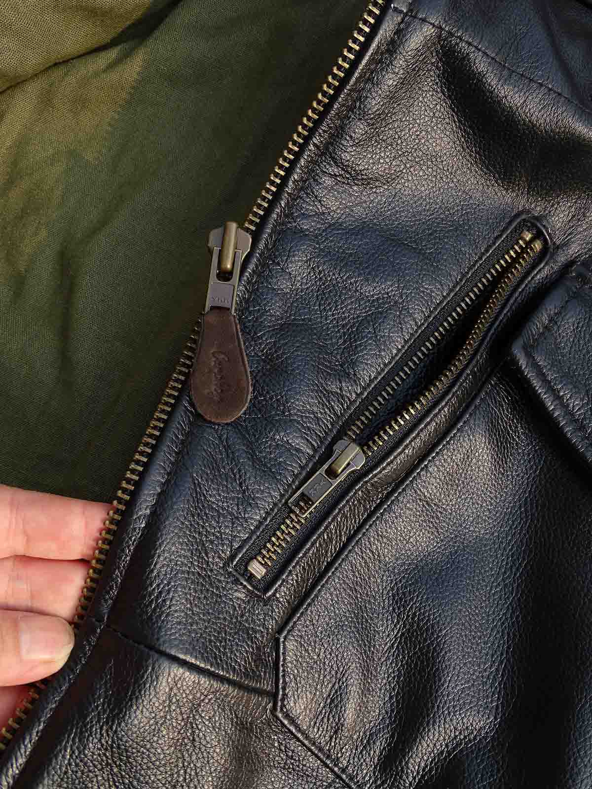 製品画像： 9ポケットフィールドフード付きフルグレインレザージャケット全体イメージ画像。黒いシボ加工レザーとヴィンテージ感漂うオイル仕上げのスエードディテールが特徴で、アウトドアやバイクライドにも適した多機能デザイン。
詳細画像： YKKアンティークブロンズジッパーとオイルドスエードレザーの補強ディテールをクローズアップした画像。耐久性とクラシックな風合いが際立つ仕上がり。