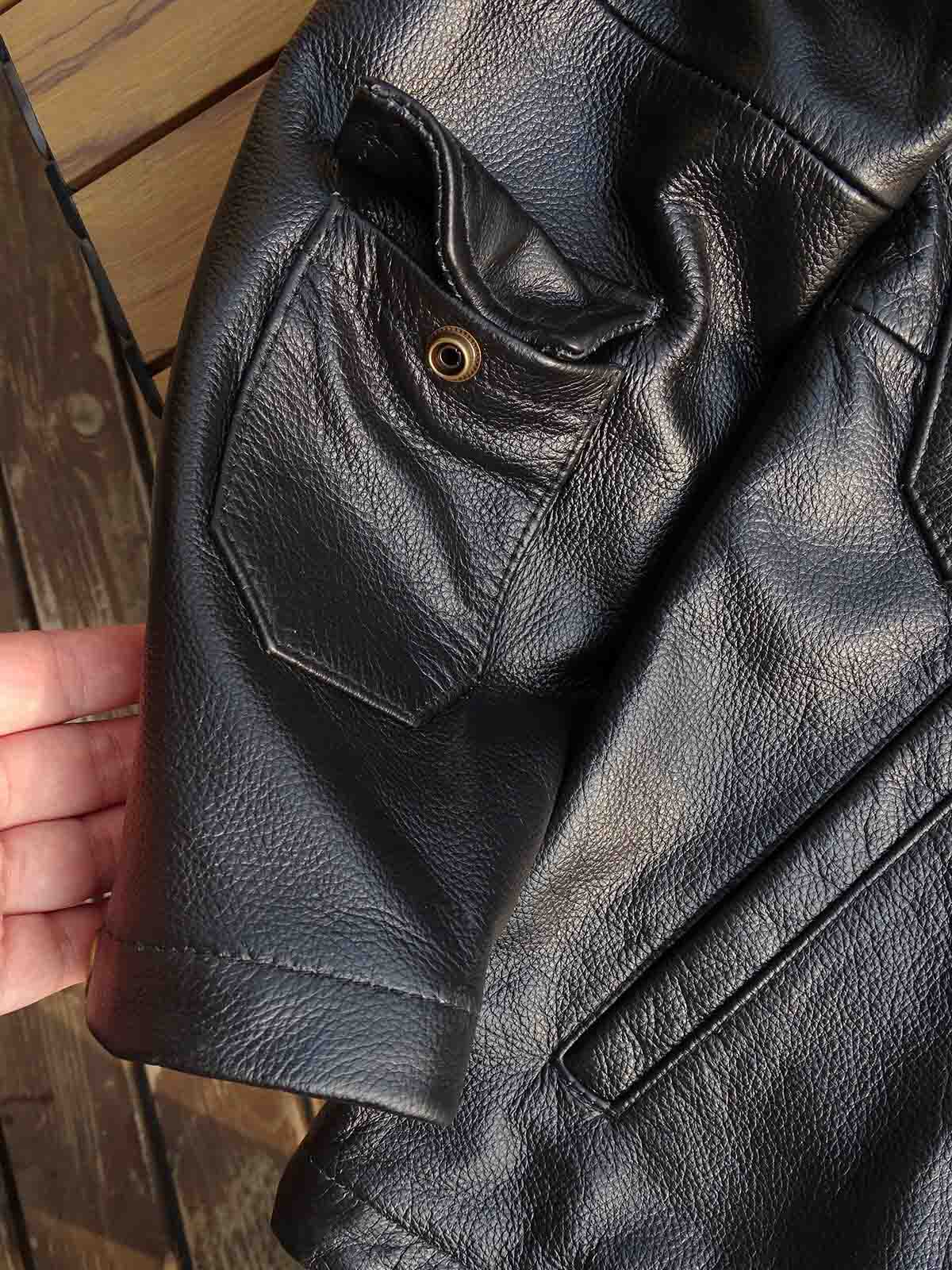 製品画像： 9ポケットフィールドフード付きフルグレインレザージャケット全体イメージ画像。黒いシボ加工レザーとヴィンテージ感漂うオイル仕上げのスエードディテールが特徴で、アウトドアやバイクライドにも適した多機能デザイン。
詳細画像： YKKアンティークブロンズジッパーとオイルドスエードレザーの補強ディテールをクローズアップした画像。耐久性とクラシックな風合いが際立つ仕上がり。