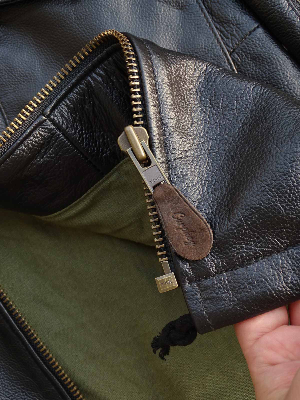 製品画像： 9ポケットフィールドフード付きフルグレインレザージャケット全体イメージ画像。黒いシボ加工レザーとヴィンテージ感漂うオイル仕上げのスエードディテールが特徴で、アウトドアやバイクライドにも適した多機能デザイン。
詳細画像： YKKアンティークブロンズジッパーとオイルドスエードレザーの補強ディテールをクローズアップした画像。耐久性とクラシックな風合いが際立つ仕上がり。