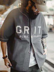 メテオライトグレーのベースボールシャツ、胸元に「GRIT PERSEVERANCE」フェルトパッチが施されている