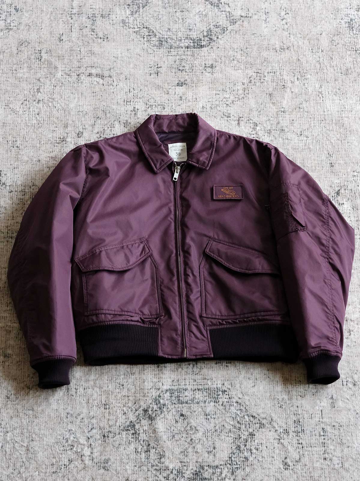 製品画像:
CWU-45Pフライトジャケットの全体展示。高密度ナイロン外層と厚手のキルティング綿を使用した、保温性と防風性に優れたクラシックなフライトジャケットの全体像。
詳細画像:
TALONメタルジッパーとマジックテープエンブレムのディテール。フラップポケットやリブ編みの袖口、裾など、ミリタリーウェア特有のデザインが強調された画像。