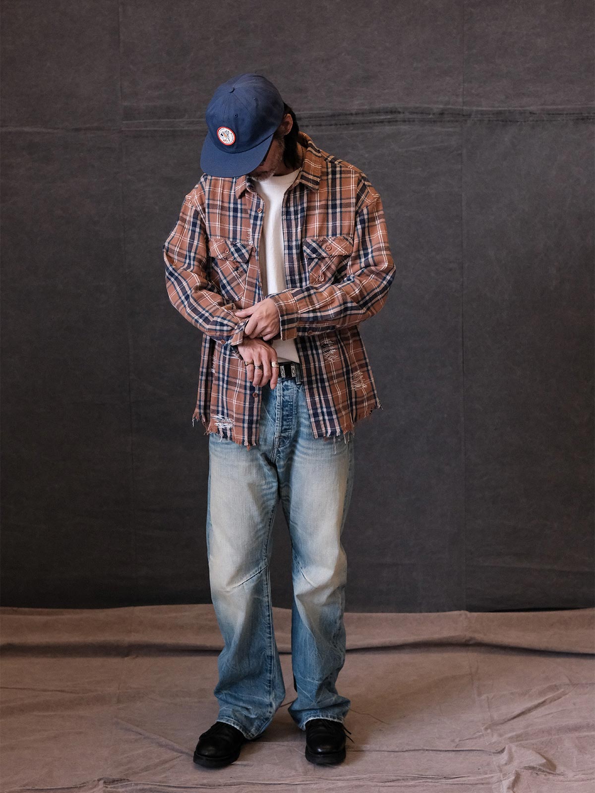 詳細画像:
ダメージ加工とチェック柄のディテール。手作業で施されたダメージデザインと、チェック柄のテクスチャーが際立つディテール。
モデル画像:
モデルがウォッシュドダメージチェックシャツを着用し、クラシックなアメリカンワークスタイルでヴィンテージコーディネートを披露している画像。