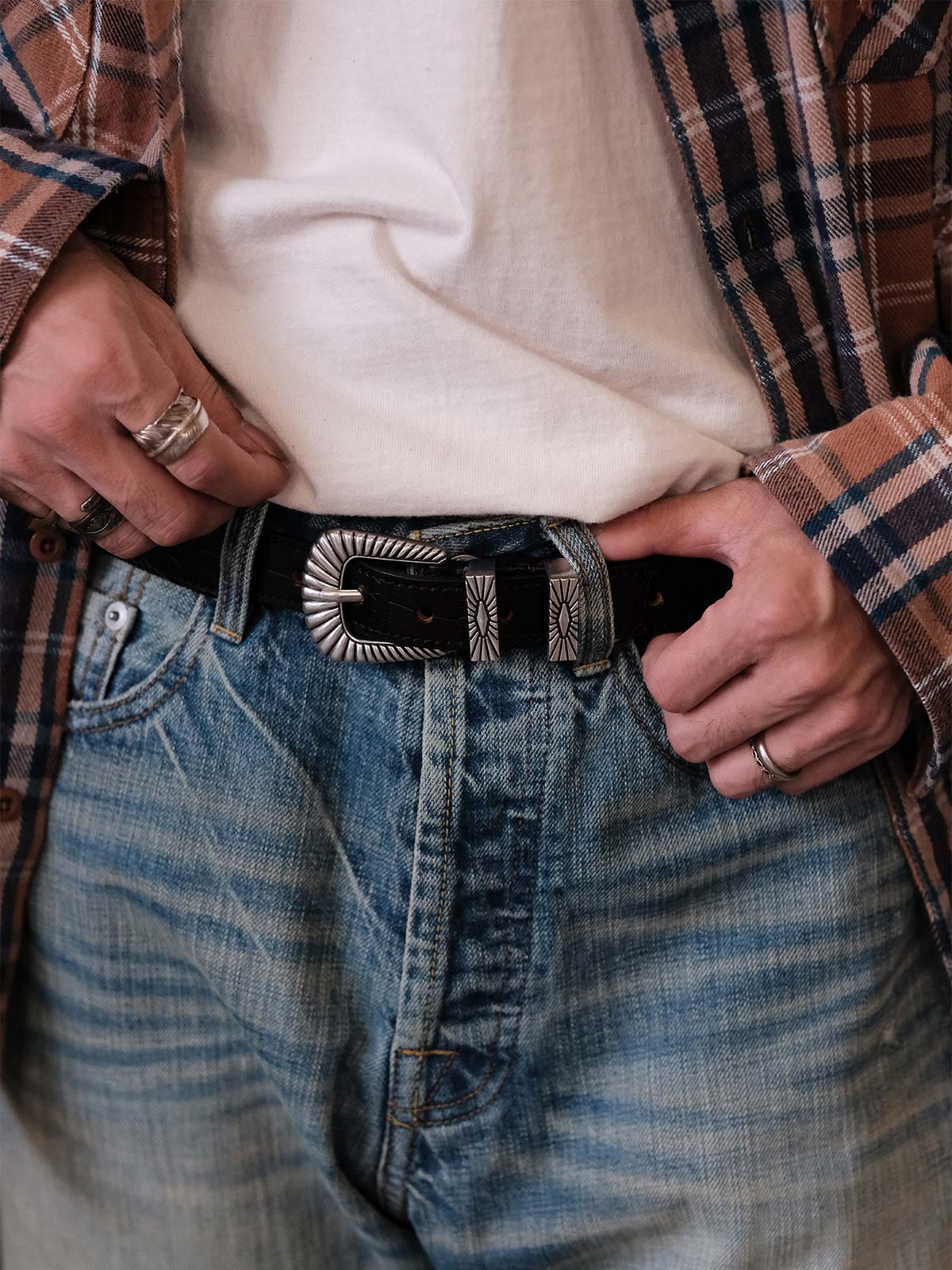 詳細画像:
エッチング加工された真鍮バックルのディテール。精緻な彫刻が施された立体感のあるバックルが、ヴィンテージスタイルの魅力を引き立てるディテール。
モデル画像:
モデルがヴィンテージキャバルリーベルトを着用し、デニムやカーゴパンツと合わせて、アメリカンウェスタンスタイルを披露している画像。
