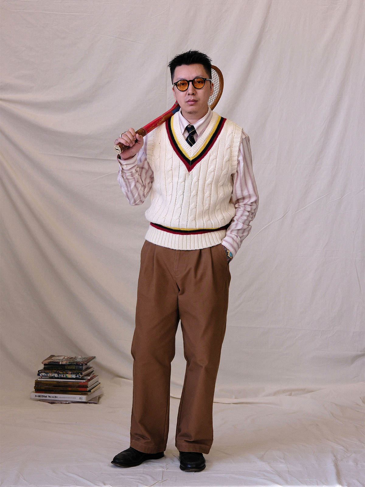 詳細画像:
ツイスト編みの編み目とカラーコンビネーションストライプのディテール。Vネックとストライプデザインが加える視覚的なレイヤード感を強調した画像。
モデル画像:
モデルがシャツと重ね着したVネック クリケットニットベストを着用し、英国風のアカデミックスタイルを演出している画像。