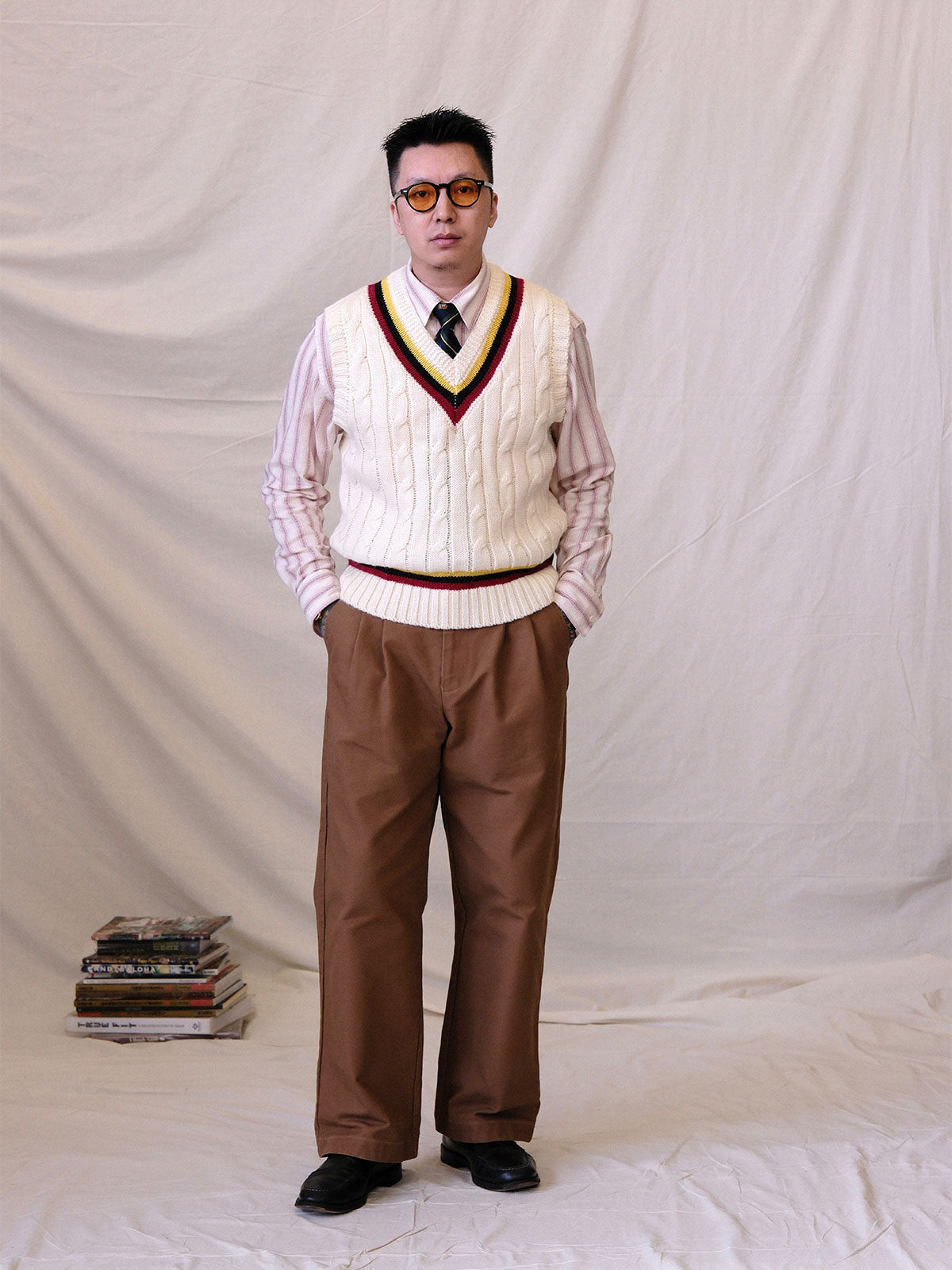 詳細画像:
ツイスト編みの編み目とカラーコンビネーションストライプのディテール。Vネックとストライプデザインが加える視覚的なレイヤード感を強調した画像。
モデル画像:
モデルがシャツと重ね着したVネック クリケットニットベストを着用し、英国風のアカデミックスタイルを演出している画像。