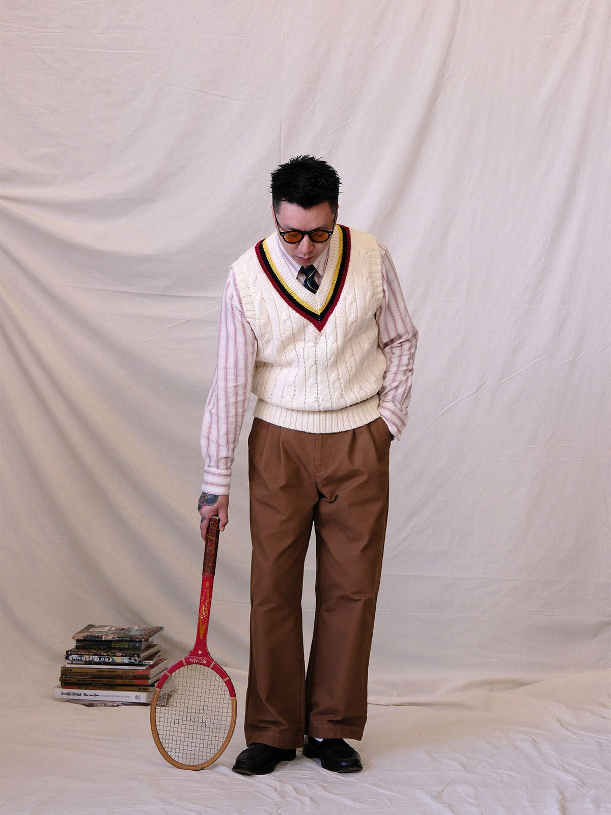 詳細画像:
ツイスト編みの編み目とカラーコンビネーションストライプのディテール。Vネックとストライプデザインが加える視覚的なレイヤード感を強調した画像。
モデル画像:
モデルがシャツと重ね着したVネック クリケットニットベストを着用し、英国風のアカデミックスタイルを演出している画像。