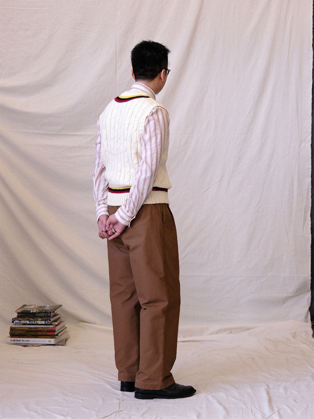 詳細画像:
ツイスト編みの編み目とカラーコンビネーションストライプのディテール。Vネックとストライプデザインが加える視覚的なレイヤード感を強調した画像。
モデル画像:
モデルがシャツと重ね着したVネック クリケットニットベストを着用し、英国風のアカデミックスタイルを演出している画像。