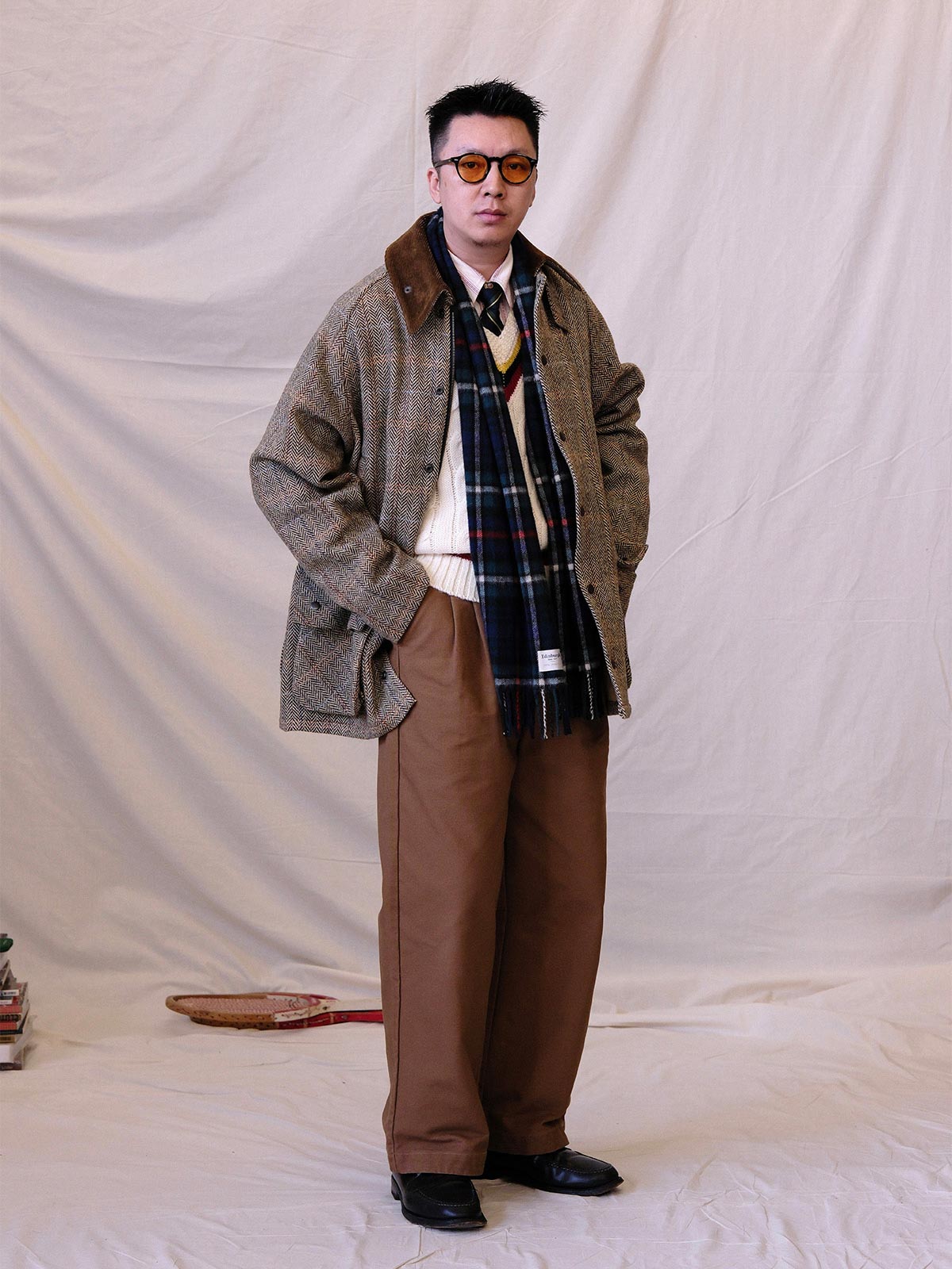 詳細画像:
ツイスト編みの編み目とカラーコンビネーションストライプのディテール。Vネックとストライプデザインが加える視覚的なレイヤード感を強調した画像。
モデル画像:
モデルがシャツと重ね着したVネック クリケットニットベストを着用し、英国風のアカデミックスタイルを演出している画像。