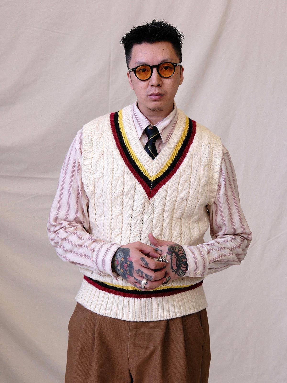 詳細画像:
ツイスト編みの編み目とカラーコンビネーションストライプのディテール。Vネックとストライプデザインが加える視覚的なレイヤード感を強調した画像。
モデル画像:
モデルがシャツと重ね着したVネック クリケットニットベストを着用し、英国風のアカデミックスタイルを演出している画像。