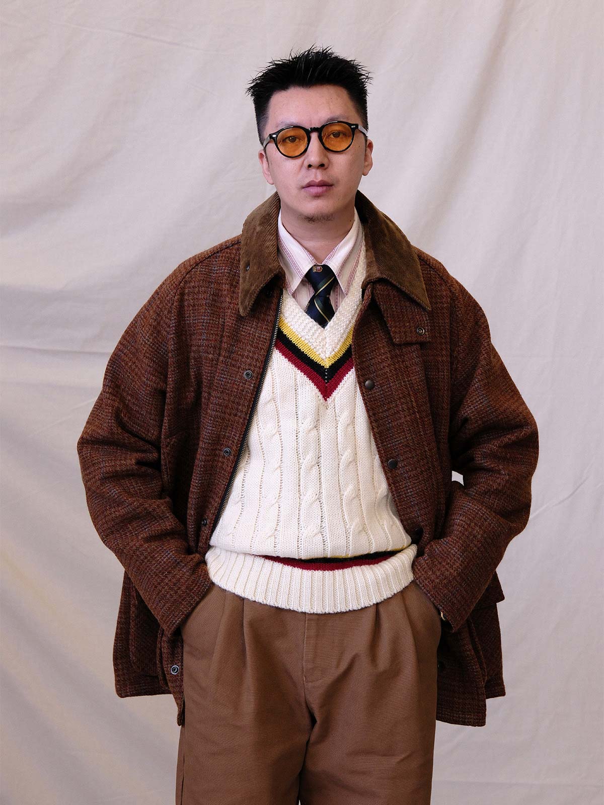 詳細画像:
ツイスト編みの編み目とカラーコンビネーションストライプのディテール。Vネックとストライプデザインが加える視覚的なレイヤード感を強調した画像。
モデル画像:
モデルがシャツと重ね着したVネック クリケットニットベストを着用し、英国風のアカデミックスタイルを演出している画像。