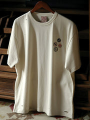 Tシャツのダメージ加工部分とデジタルプリントされたピンバッジのクローズアップ。先進的なプリント技術とヴィンテージ感を演出する手作業の加工が見られます。