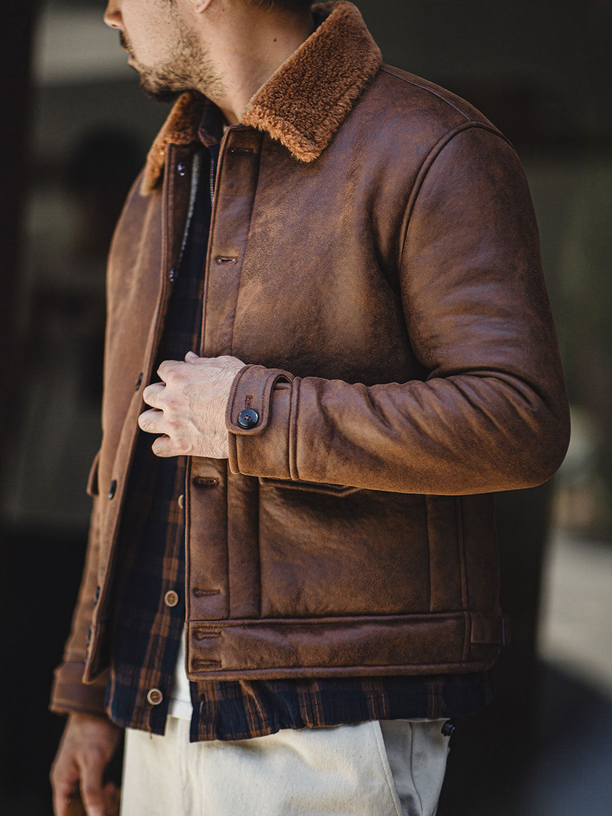 詳細画像：七つボタンの前立てと厚手のシープスキン襟のディテールをクローズアップ。ヴィンテージ感と実用性を兼ね備えた仕様。
モデル画像：モデルがムートンボマージャケットを着用し、デニムパンツとワークブーツを合わせたハードなバイカースタイルを提案。