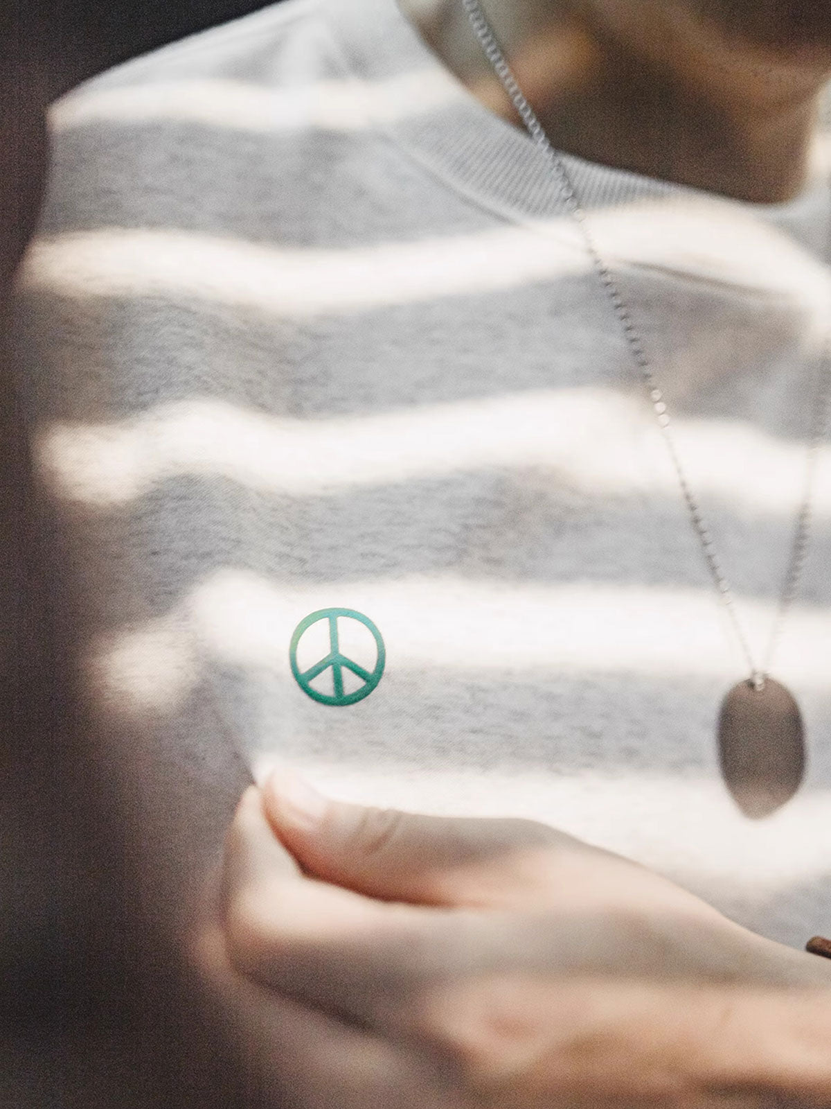 製品画像： ライトグレーの裏起毛クルーネックスウェット全体イメージ画像。リラックスシルエットとドロップショルダーデザインが特徴で、胸元には「ピースマーク」プリントがアクセント。単独でもインナーとしても活躍する秋冬の万能アイテム。
詳細画像： 胸元に配置された精巧な「ピースマーク」プリントとクラシックなリブ仕様のネックや裾をクローズアップした画像。保温性と耐久性を兼ね備えた裏起毛素材が際立つ。
