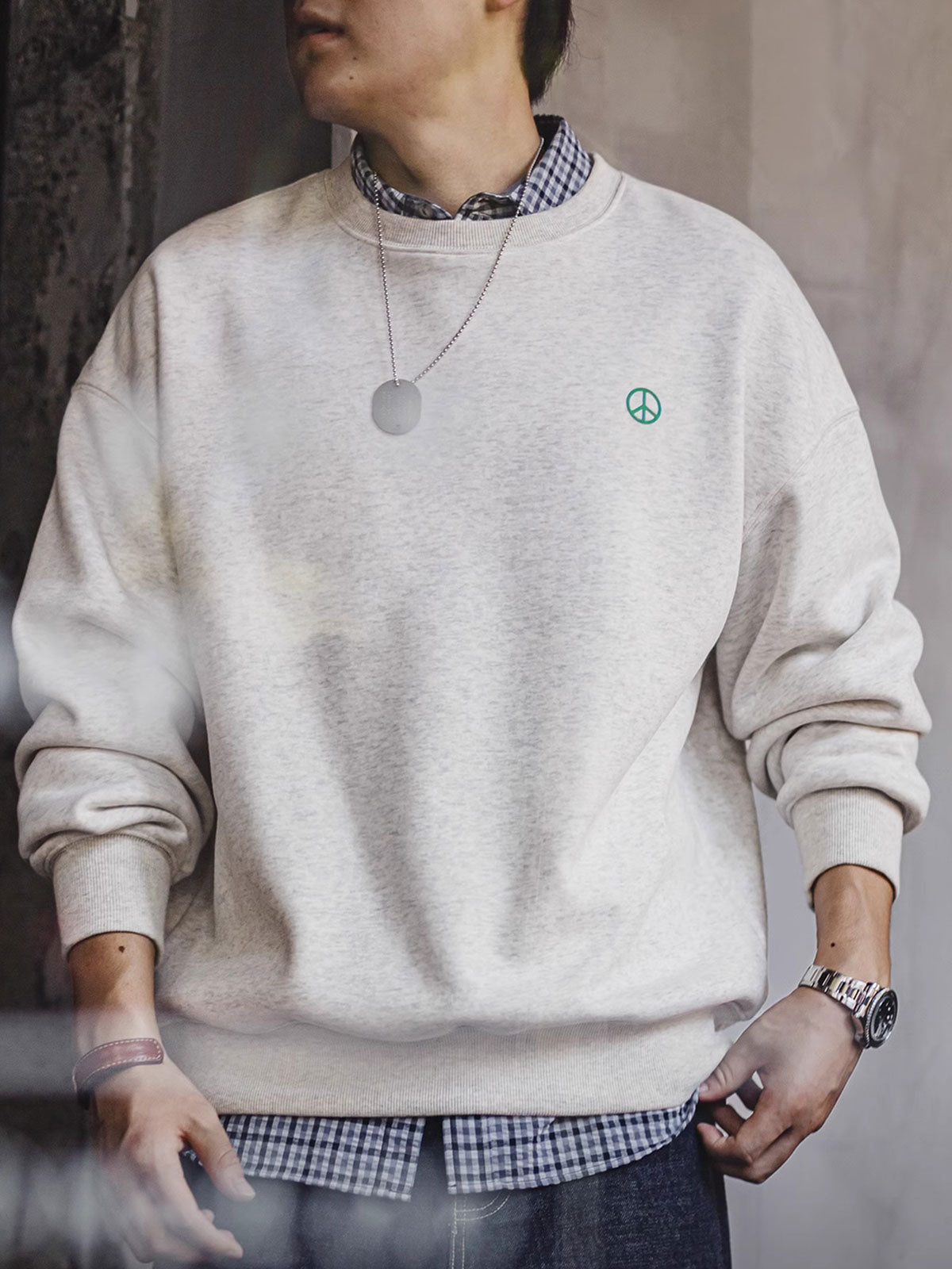詳細画像： 胸元に配置された精巧な「ピースマーク」プリントとクラシックなリブ仕様のネックや裾をクローズアップした画像。保温性と耐久性を兼ね備えた裏起毛素材が際立つ。
モデル画像： 裏起毛クルーネックスウェットを着用したモデルが、スニーカーとブラックパンツを合わせたカジュアルな秋冬スタイルを提案。快適さと個性を両立したコーディネートが魅力的。
