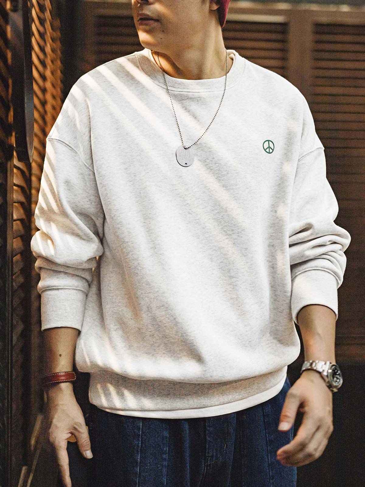詳細画像： 胸元に配置された精巧な「ピースマーク」プリントとクラシックなリブ仕様のネックや裾をクローズアップした画像。保温性と耐久性を兼ね備えた裏起毛素材が際立つ。
モデル画像： 裏起毛クルーネックスウェットを着用したモデルが、スニーカーとブラックパンツを合わせたカジュアルな秋冬スタイルを提案。快適さと個性を両立したコーディネートが魅力的。