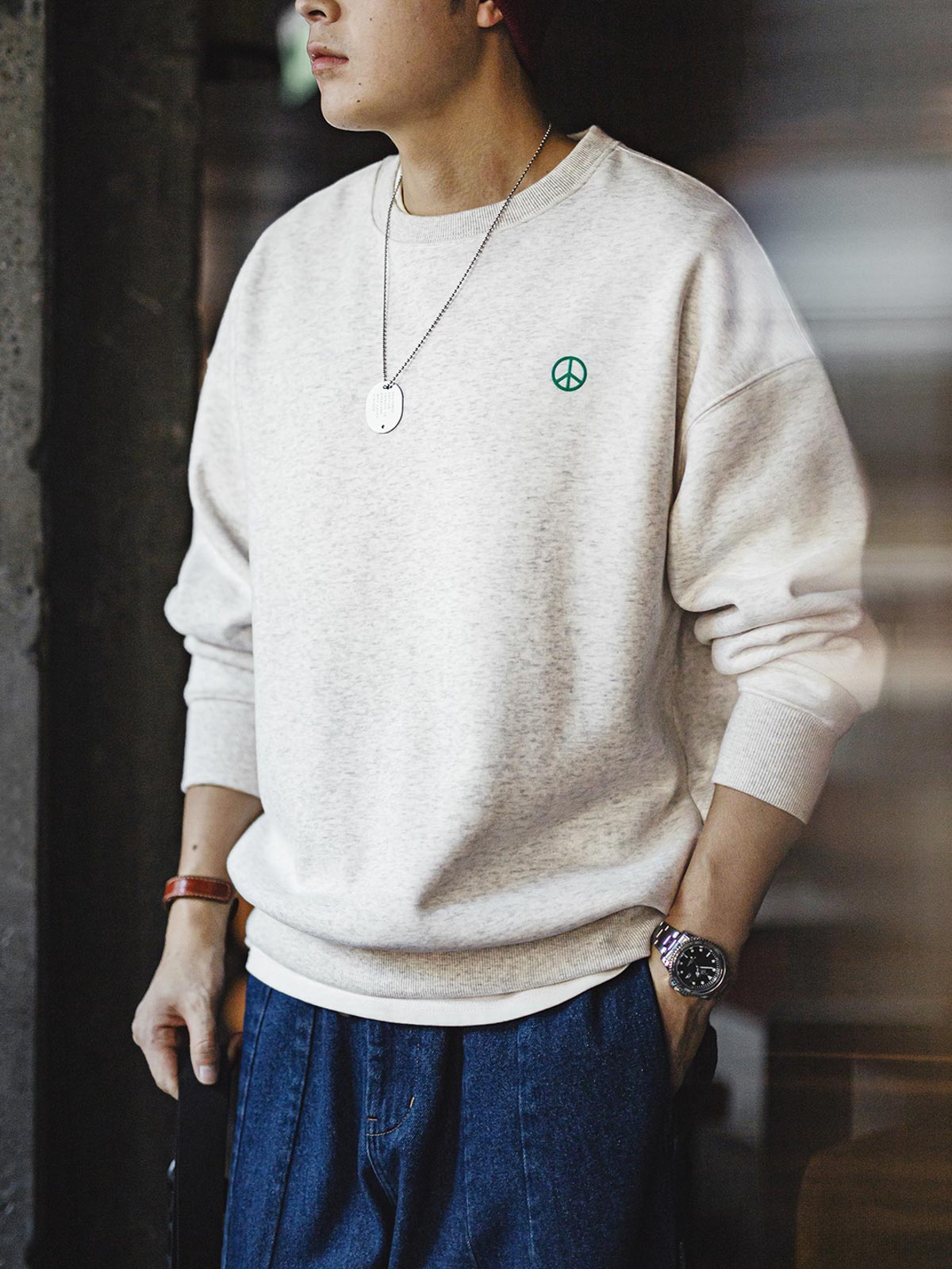 詳細画像： 胸元に配置された精巧な「ピースマーク」プリントとクラシックなリブ仕様のネックや裾をクローズアップした画像。保温性と耐久性を兼ね備えた裏起毛素材が際立つ。
モデル画像： 裏起毛クルーネックスウェットを着用したモデルが、スニーカーとブラックパンツを合わせたカジュアルな秋冬スタイルを提案。快適さと個性を両立したコーディネートが魅力的。