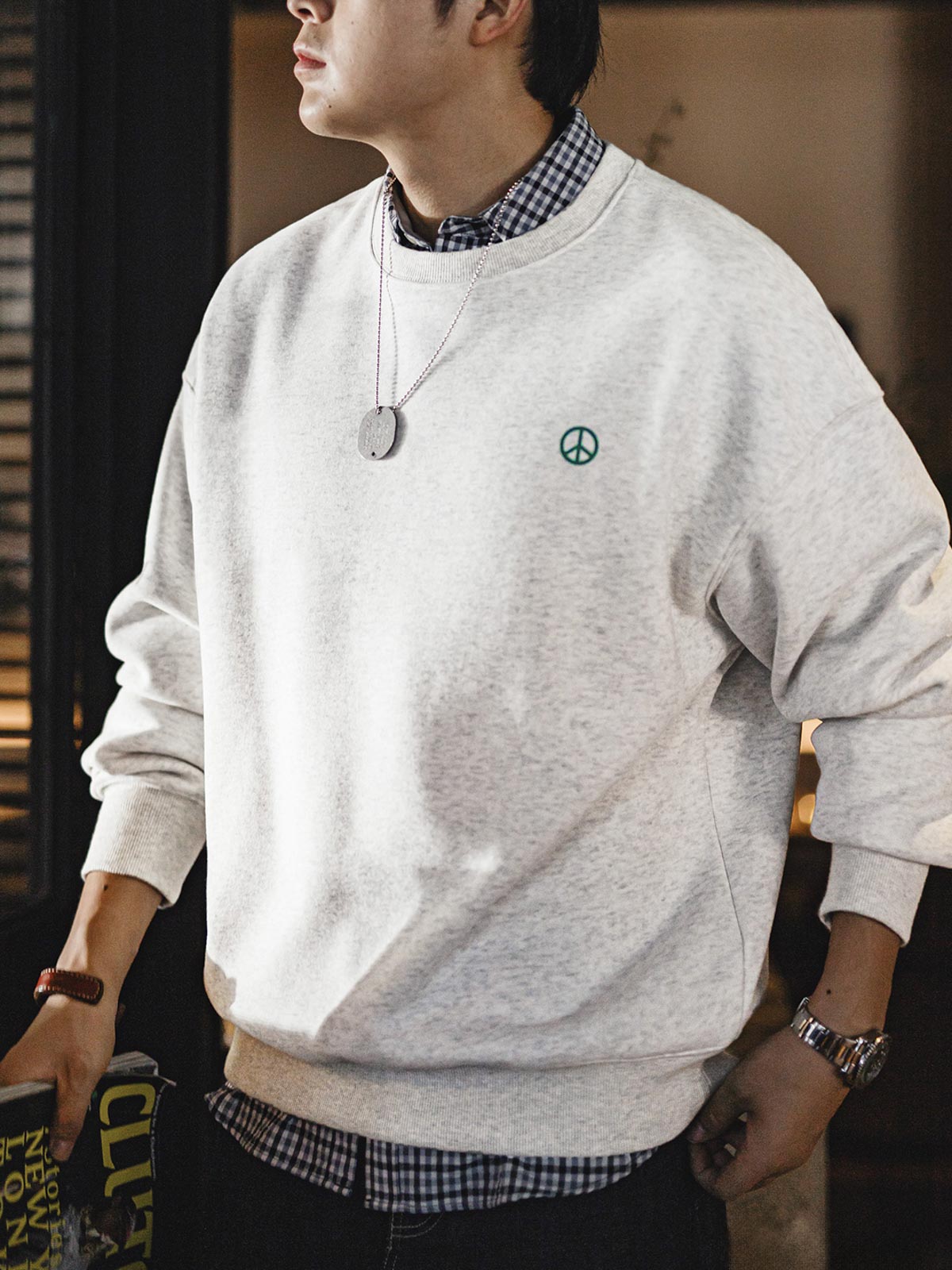 詳細画像： 胸元に配置された精巧な「ピースマーク」プリントとクラシックなリブ仕様のネックや裾をクローズアップした画像。保温性と耐久性を兼ね備えた裏起毛素材が際立つ。
モデル画像： 裏起毛クルーネックスウェットを着用したモデルが、スニーカーとブラックパンツを合わせたカジュアルな秋冬スタイルを提案。快適さと個性を両立したコーディネートが魅力的。