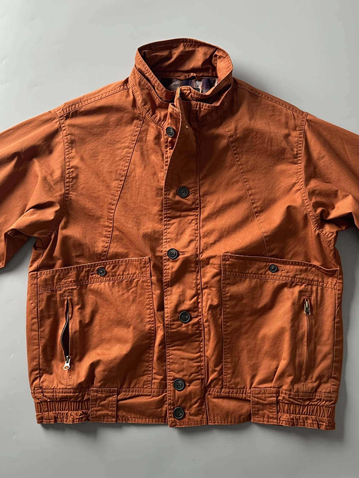 製品画像: オレンジカラーのダブルスタンドカラーワークジャケットの正面画像、実用的でスタイリッシュなデザインが特徴。
詳細画像: 袖部のジッパーポケットとスタンドカラーデザインのディテール画像、ワークスタイルを強調。