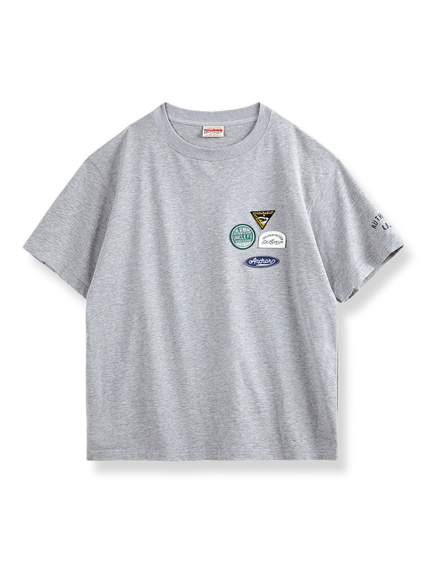 左胸に4つのカラフルな刺繍バッジが付いたグレーベースの半袖Tシャツ。
