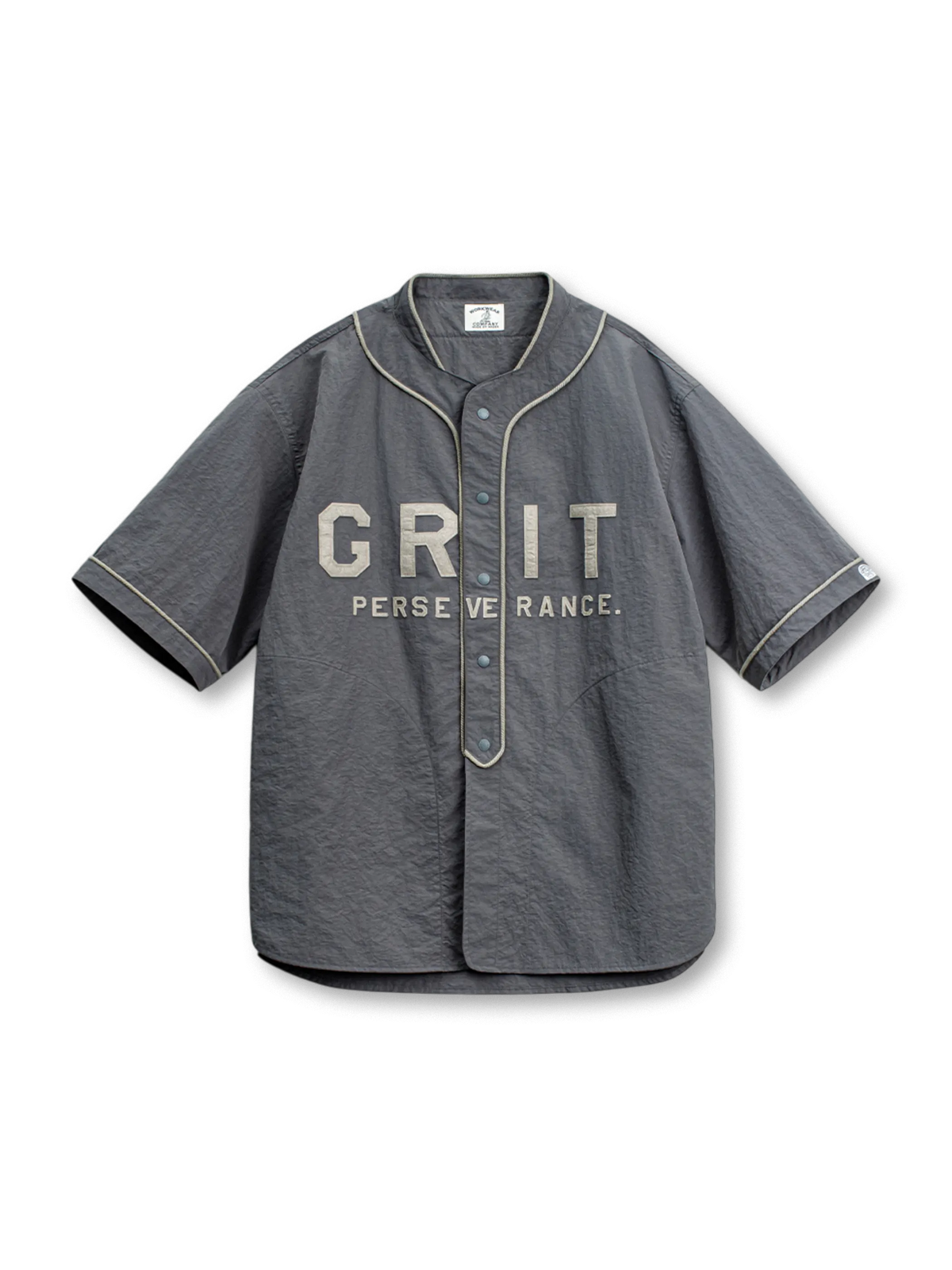 「GRIT PERSEVERANCE」パッチとホログラムボタンが特徴の90年代風軽量ベースボールシャツ。