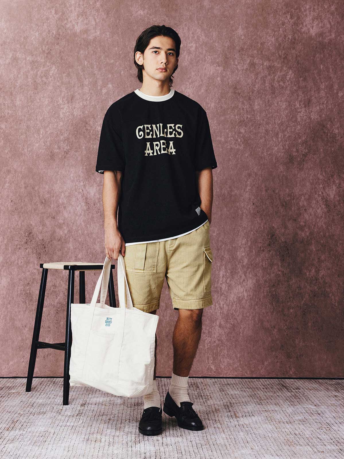 モデルがフットボールカルチャープリントTシャツを着用し、ジンジャーイエローのバリエーションで、ダークカラーのカジュアルパンツとスポーツシューズを合わせたカジュアルなスタイリングでポーズをとる画像。レトロかつ現代的な夏の装いを演出。